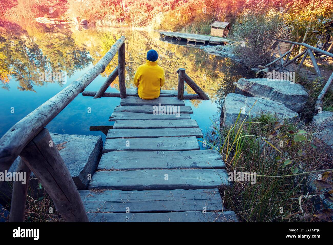 Un homme assis sur une terrasse en bois et regardant un lac forestier avec une rive en granit. Lever du soleil sur le lac en automne Banque D'Images
