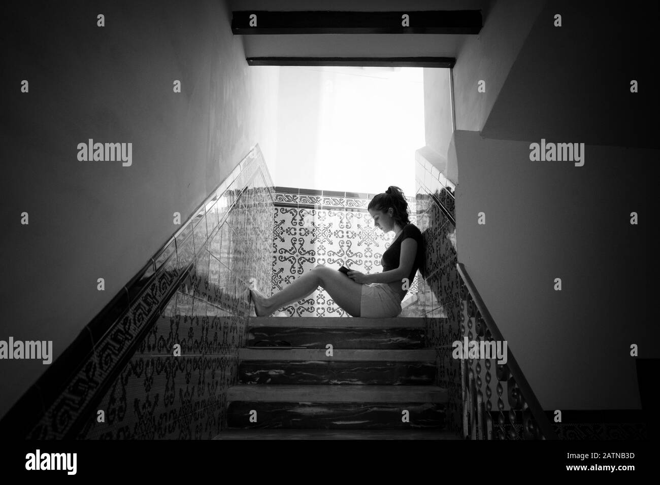 Jeune femme assise sur les escaliers d'une ancienne maison méditerranéenne traditionnelle lecture d'un livre avec lumière naturelle venant de derrière. B&O. Noir et blanc Banque D'Images