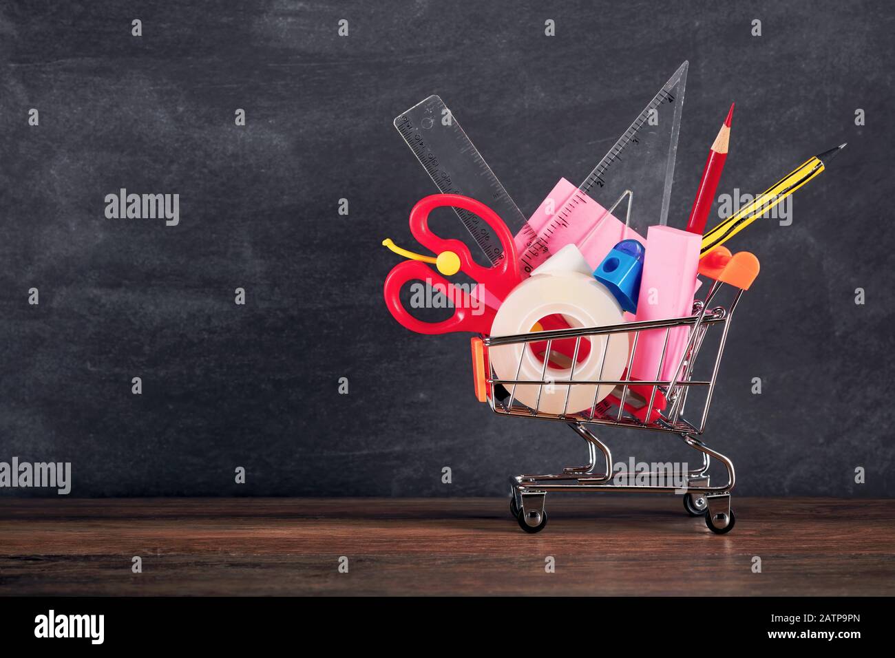 Articles de papeterie dans un panier d'achat sur un bureau en bois contre le tableau noir. Retour au concept scolaire. Banque D'Images