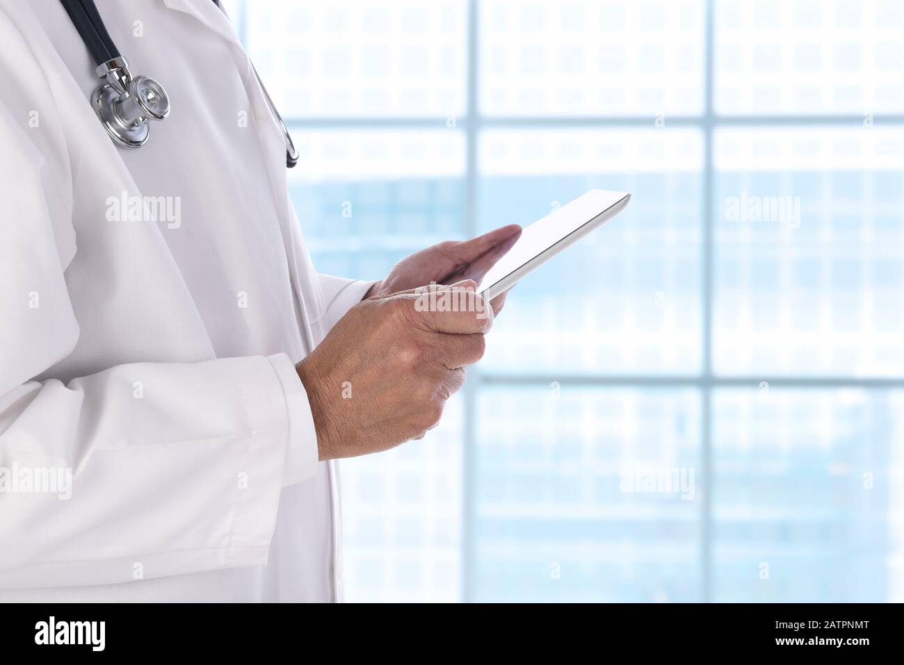 Gros plan d'un professionnel de la santé tenant une tablette informatique tout en se tenant devant une grande fenêtre dans un établissement médical moderne. Banque D'Images