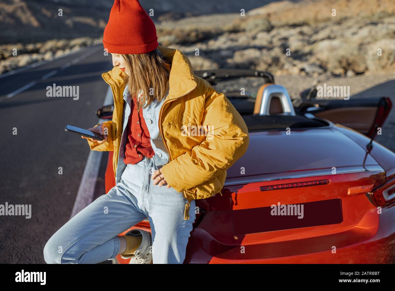 Femme élégante, veste jaune et chapeau rouge, en voyage, assise avec un  smartphone sur le capot de la voiture, sur le bord de la vallée du désert,  au coucher du soleil Photo
