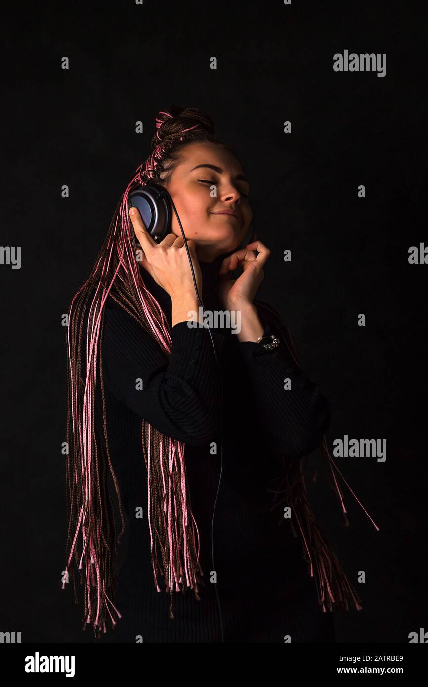 Belle fille avec des piquets africains zizi écoute avec des yeux fermés la musique dans des écouteurs sur un fond noir. Émotions de bonheur, de plaisir et p Banque D'Images