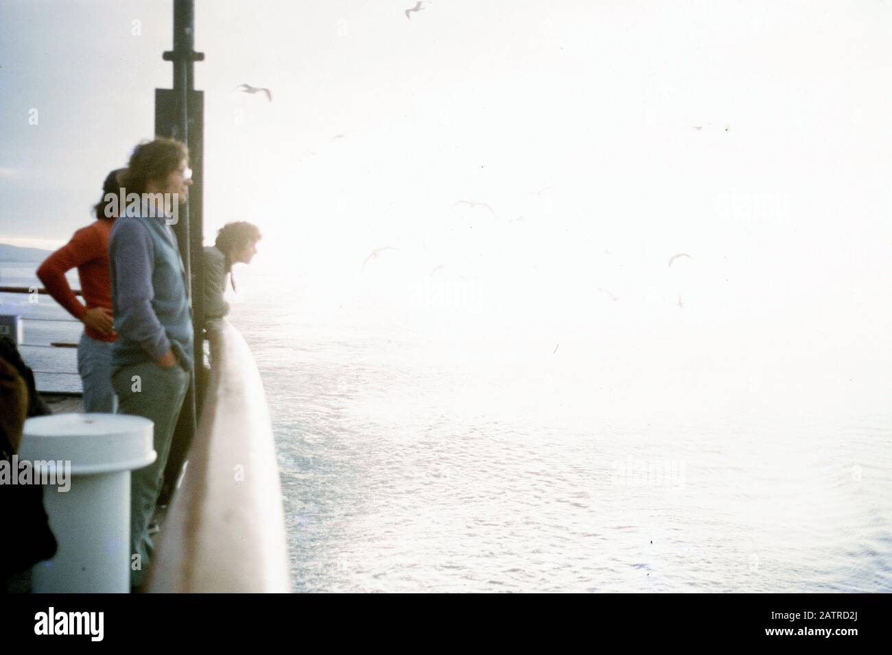 Photographie vernaculaire prise sur un film analogique de 35 mm transparent, montrant plusieurs personnes debout sur le rail du navire et donnant sur la mer, avec un vaste espace de copie à droite du cadre, suggérant la contemplation ou la méditation, 1970. () Banque D'Images