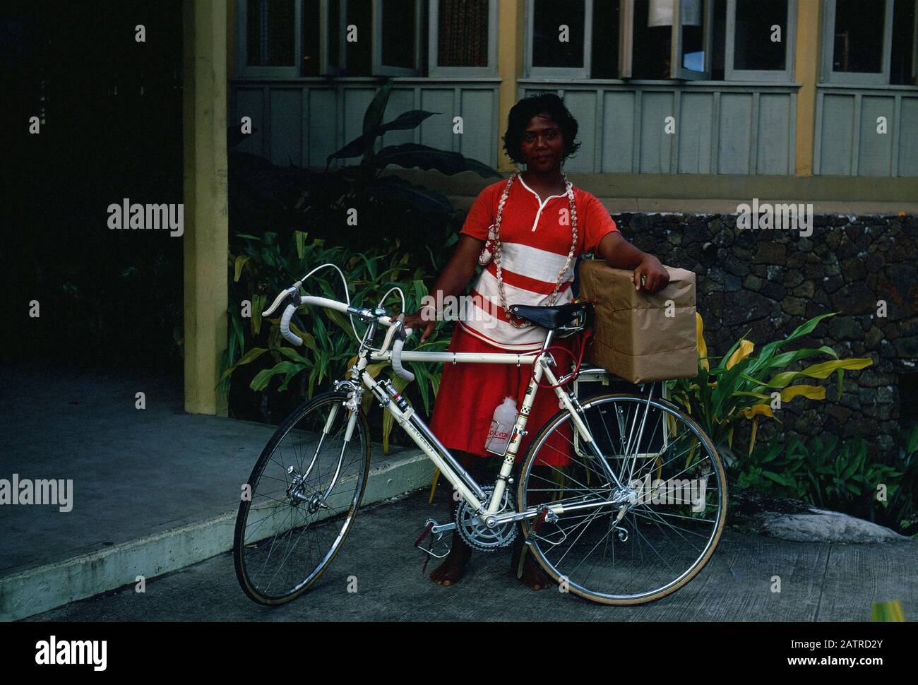 Photographie vernaculaire prise sur un film analogique de 35 mm transparent, considérée comme une femme en robe rouge et blanche à vélo rouge, 1970. Les principaux sujets/objets détectés incluent vélo et véhicule. () Banque D'Images