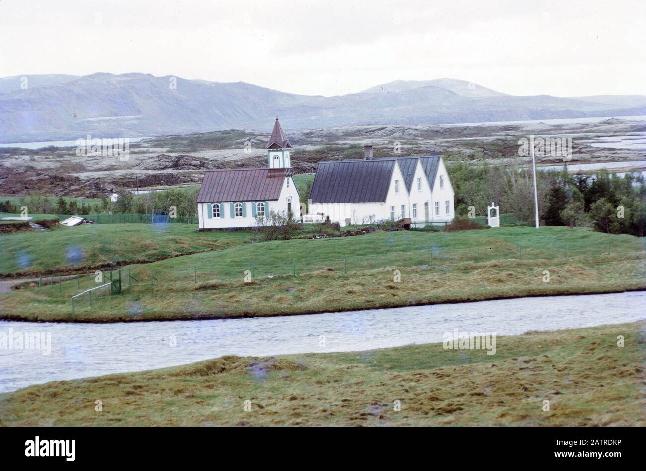 Photographie vernaculaire prise sur un film analogique de 35 mm transparent, considérée comme représentant une maison blanche et grise près du lac et du champ vert d'herbe, 1970. () Banque D'Images