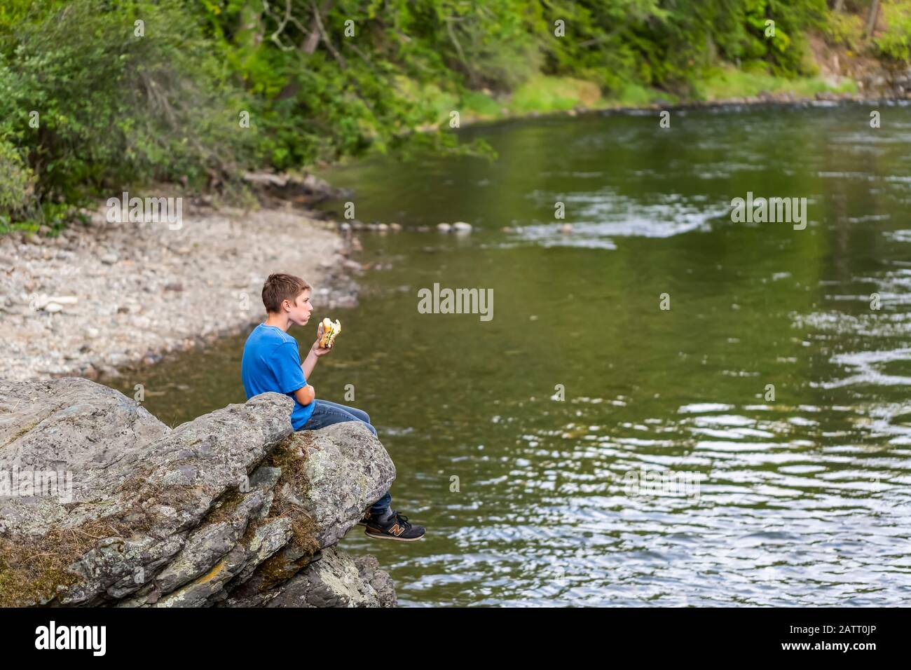 Un adolescent est assis sur une corniche au-dessus d'une rivière en train de manger un sandwich pour le déjeuner; Colombie-Britannique, Canada Banque D'Images