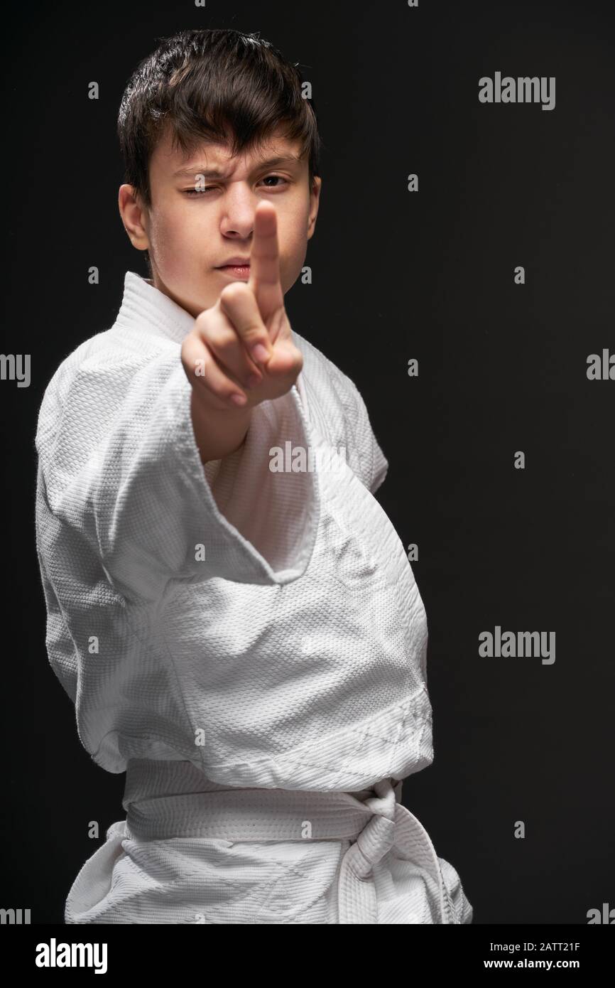 un adolescent vêtu de vêtements d'arts martiaux pointant le doigt sur un fond gris foncé, un concept sportif Banque D'Images