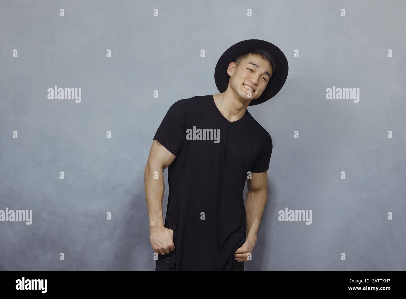 Bel homme asiatique avec coupe courte se posant dans un chapeau noir  tendance et un t-shirt noir sur fond gris dans le studio. Photo intérieure  d'un gars confiant dans un élégant vêtement
