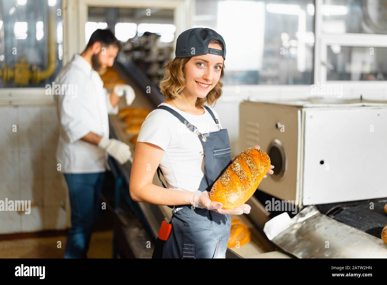 Portrait d'une jeune fille boulangère avec du pain dans les mains sur le fond d'une ligne automatisée dans une boulangerie. Production industrielle de pain Banque D'Images
