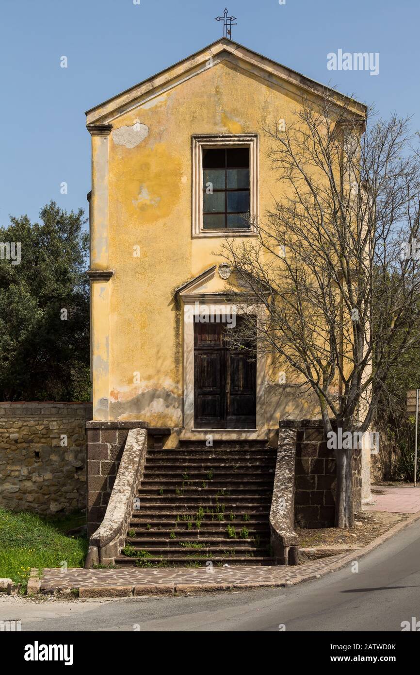 Façade jaune d'une petite église située à l'angle des rues, surmontée d'arbres et de buissons. Staris à la porte d'entrée. Traversez le toit. SanT'Orso Banque D'Images