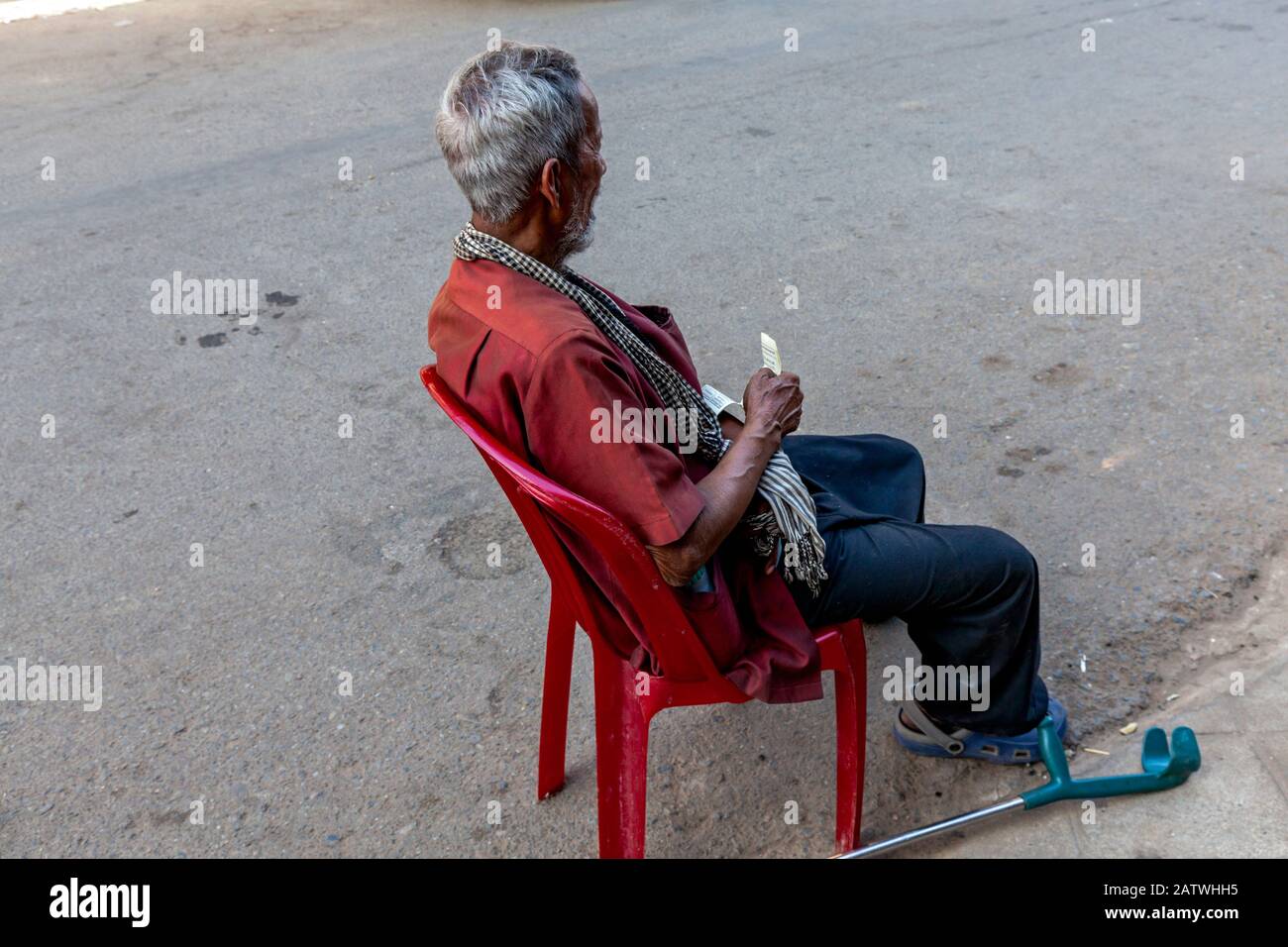 Un pauvre homme asiatique de 75 ans victime d'un accident vasculaire cérébral est assis dans une chaise en plastique rouge dans une rue de la ville dans la ville de Kampong Cham, au Cambodge. Banque D'Images