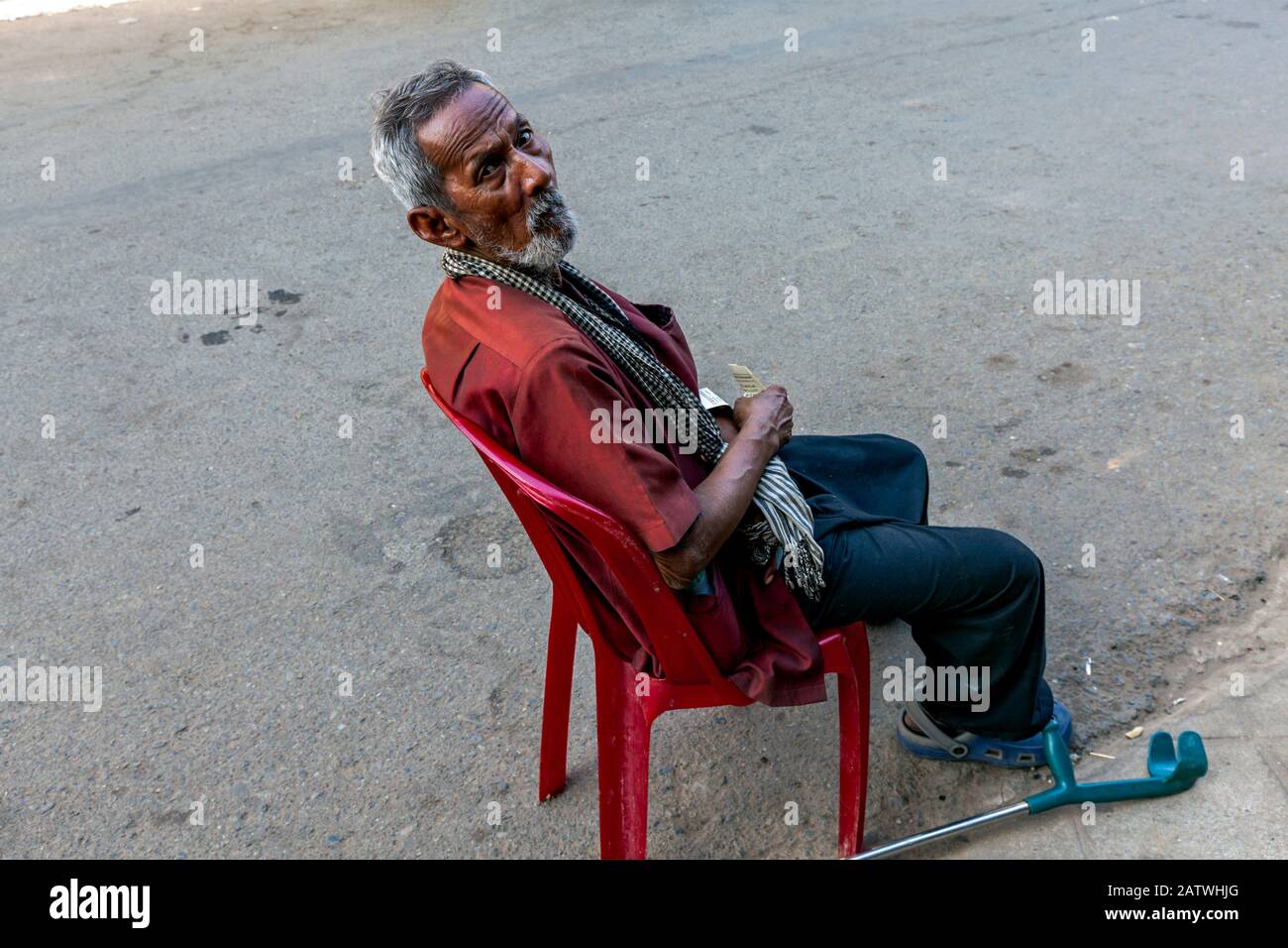Un pauvre homme asiatique de 75 ans victime d'un accident vasculaire cérébral est assis dans une chaise en plastique rouge dans une rue de la ville dans la ville de Kampong Cham, au Cambodge. Banque D'Images