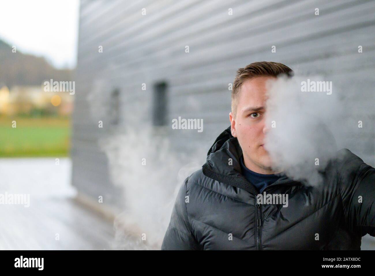 Jeune homme dans ses années vingt accro à fumer debout à l'extérieur vaporing sur une cigarette électronique soufflant un nuage de fumée vers l'appareil photo avec espace de copie Banque D'Images