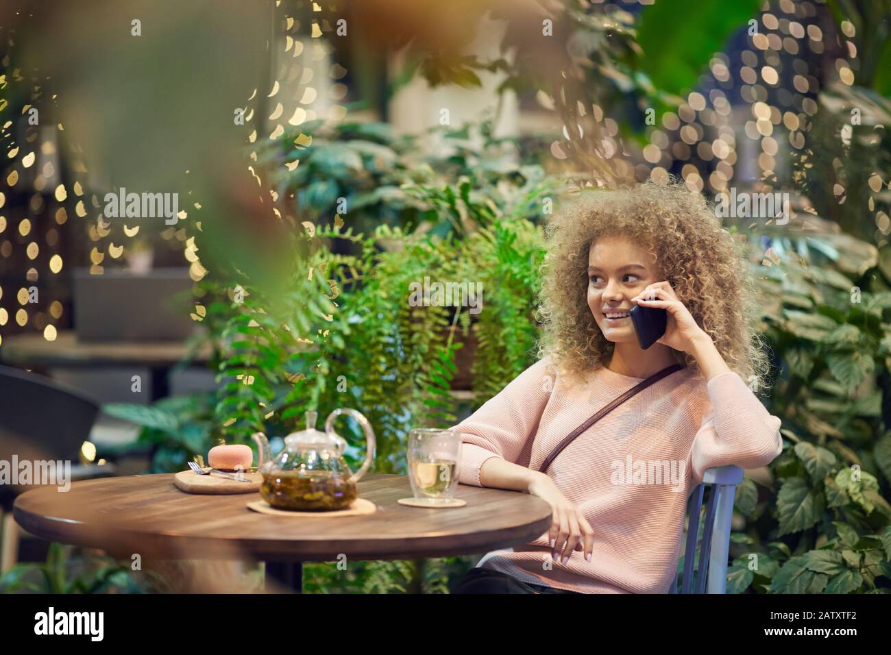 Jeune femme souriante avec des cheveux frisés se reposant dans un café avec une tasse de thé et parler sur le téléphone mobile Banque D'Images