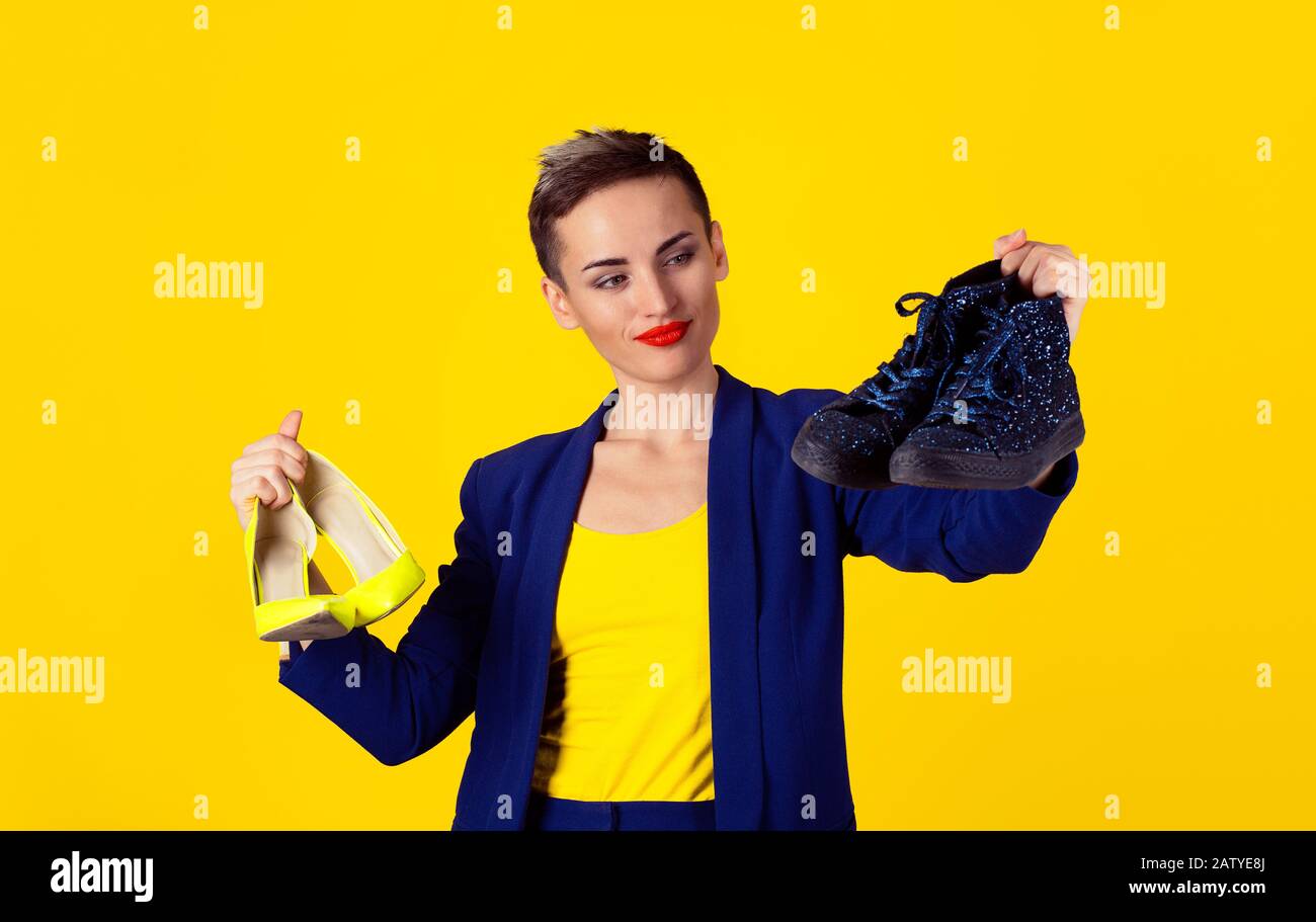 Concept de chaussures confortables. Femme cheveux court fille tenant jaune talons hauts chaussure couleur citron dans une main regardant les chaussures plates sport bleu Banque D'Images