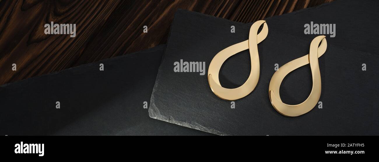 Vue de dessus des boucles d'oreilles dorées en forme d'infini sur les plaques noires sur fond en bois brun Banque D'Images