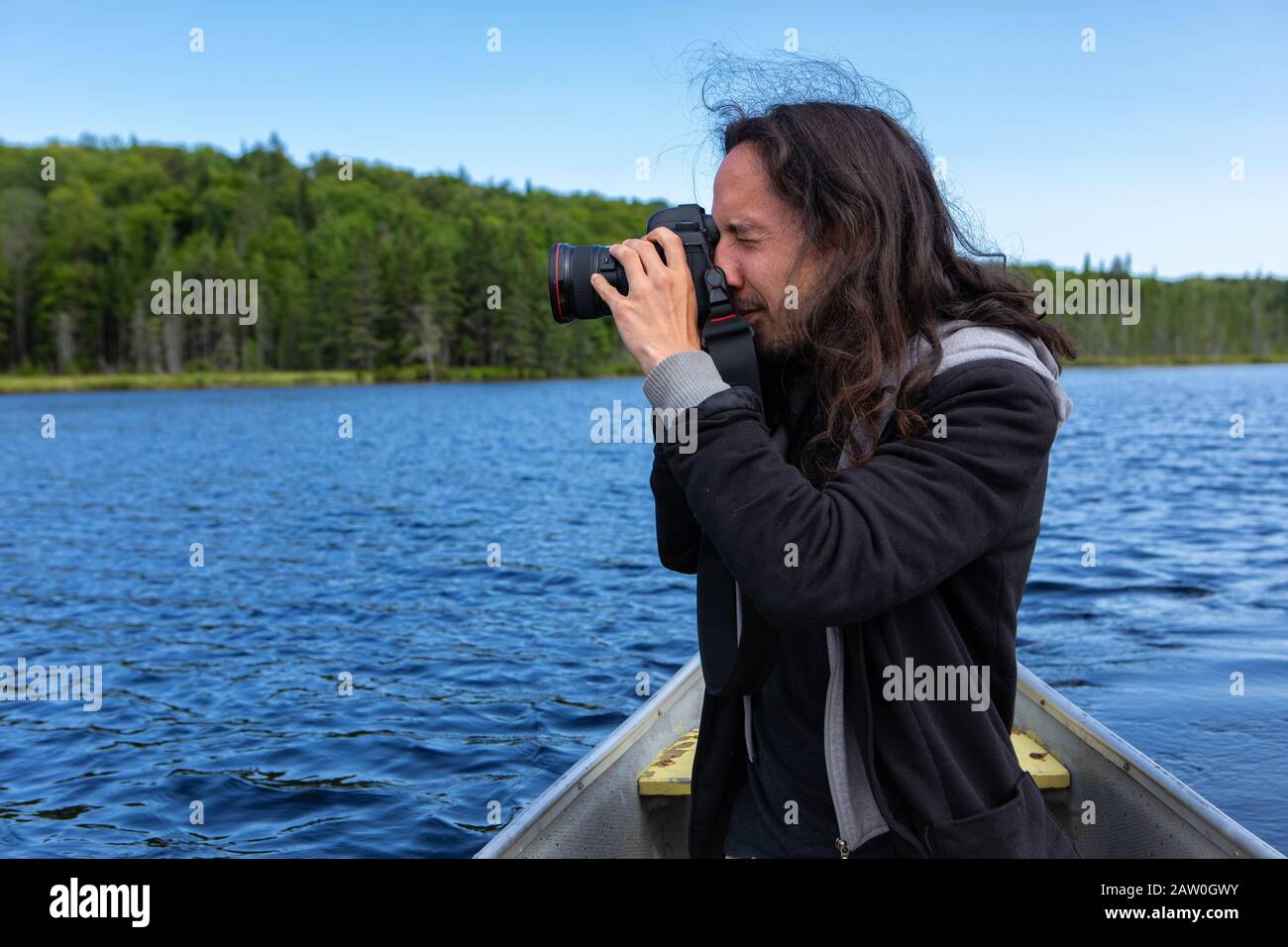 Vue latérale d'un jeune homme d'amérique indigène à poil long qui monte en canoë dans le lac tout en photographiant un paysage pittoresque dans le nord du Québec, au Canada Banque D'Images