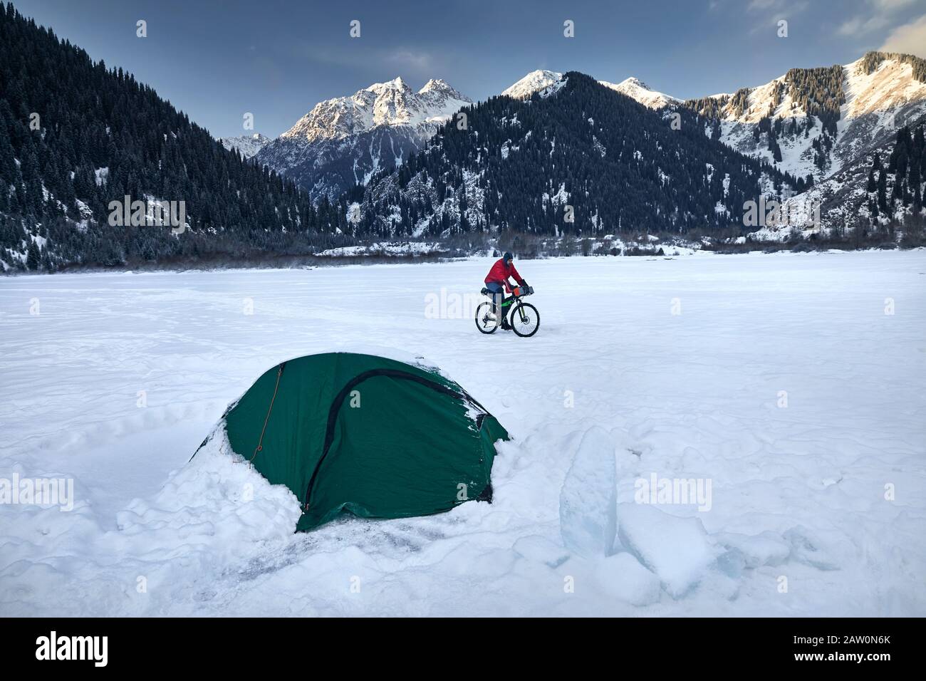 L'homme en veste rouge monte sa bicyclette au lac gelé dans les montagnes près de la tente verte au lever du soleil. Banque D'Images