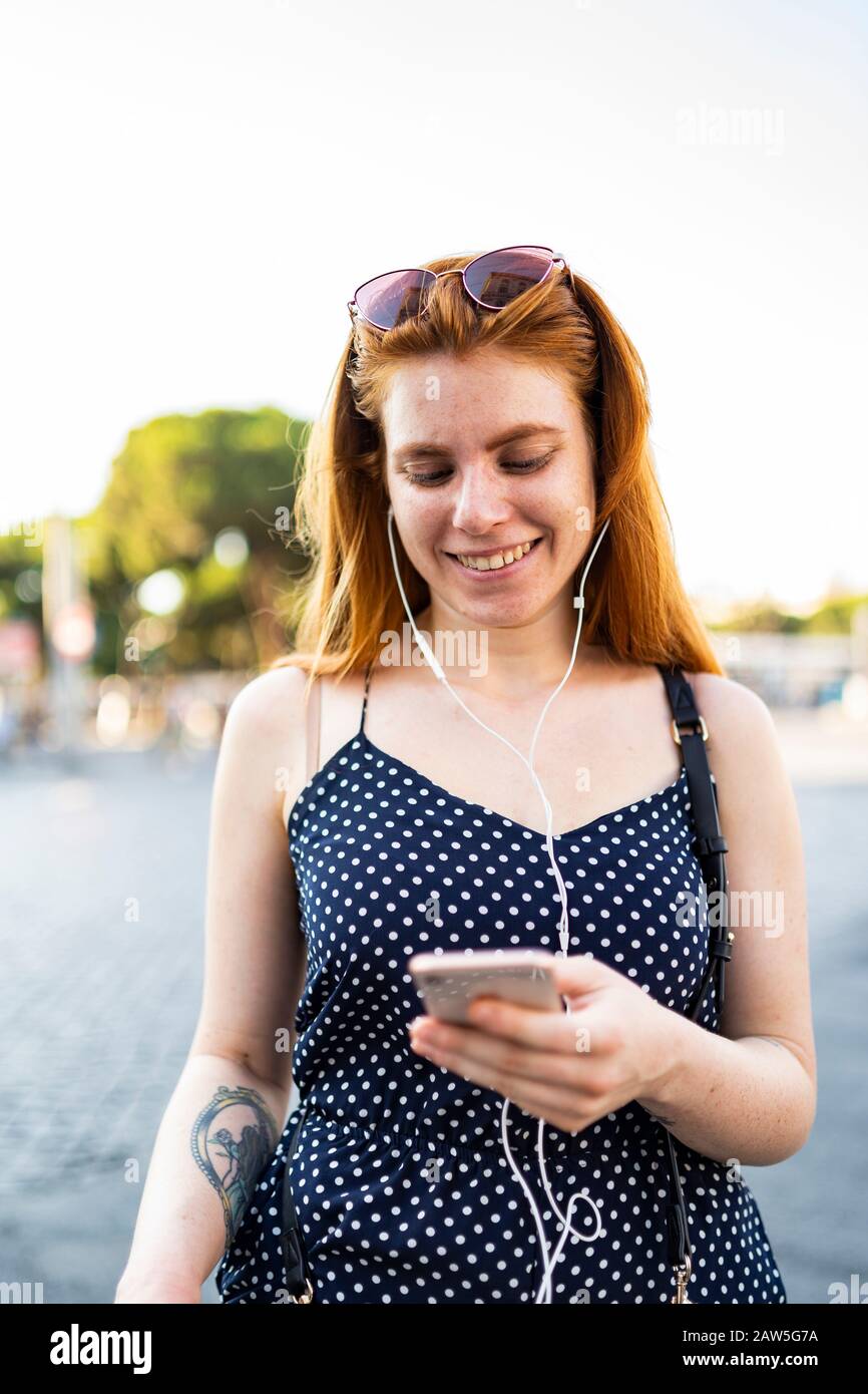 Jeune femme gaie avec cheveux rouges écoutant de la musique dans des écouteurs et lisant des messages sur smartphone tout en se tenant dans la rue le jour ensoleillé de l'été Banque D'Images