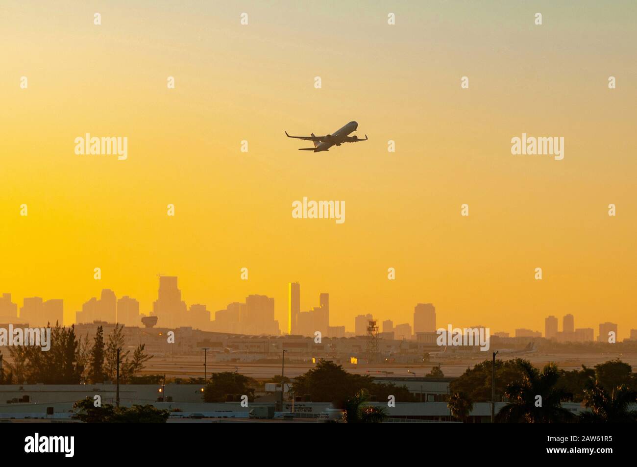 Un avion est silhouetté contre un lever de soleil coloré jaune et orange lorsqu'il part de l'aéroport de Miami avec la ville en arrière-plan. Banque D'Images