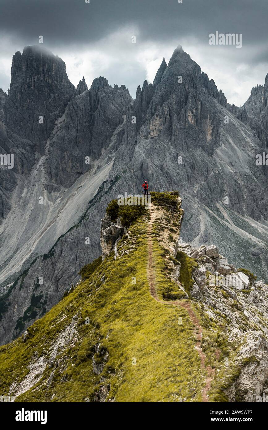 Homme avec veste rouge debout sur un degré, derrière lui des pics de montagne et des roches pointues, des nuages dramatiques, Cimon le groupe Croda Liscia et Cadini Banque D'Images