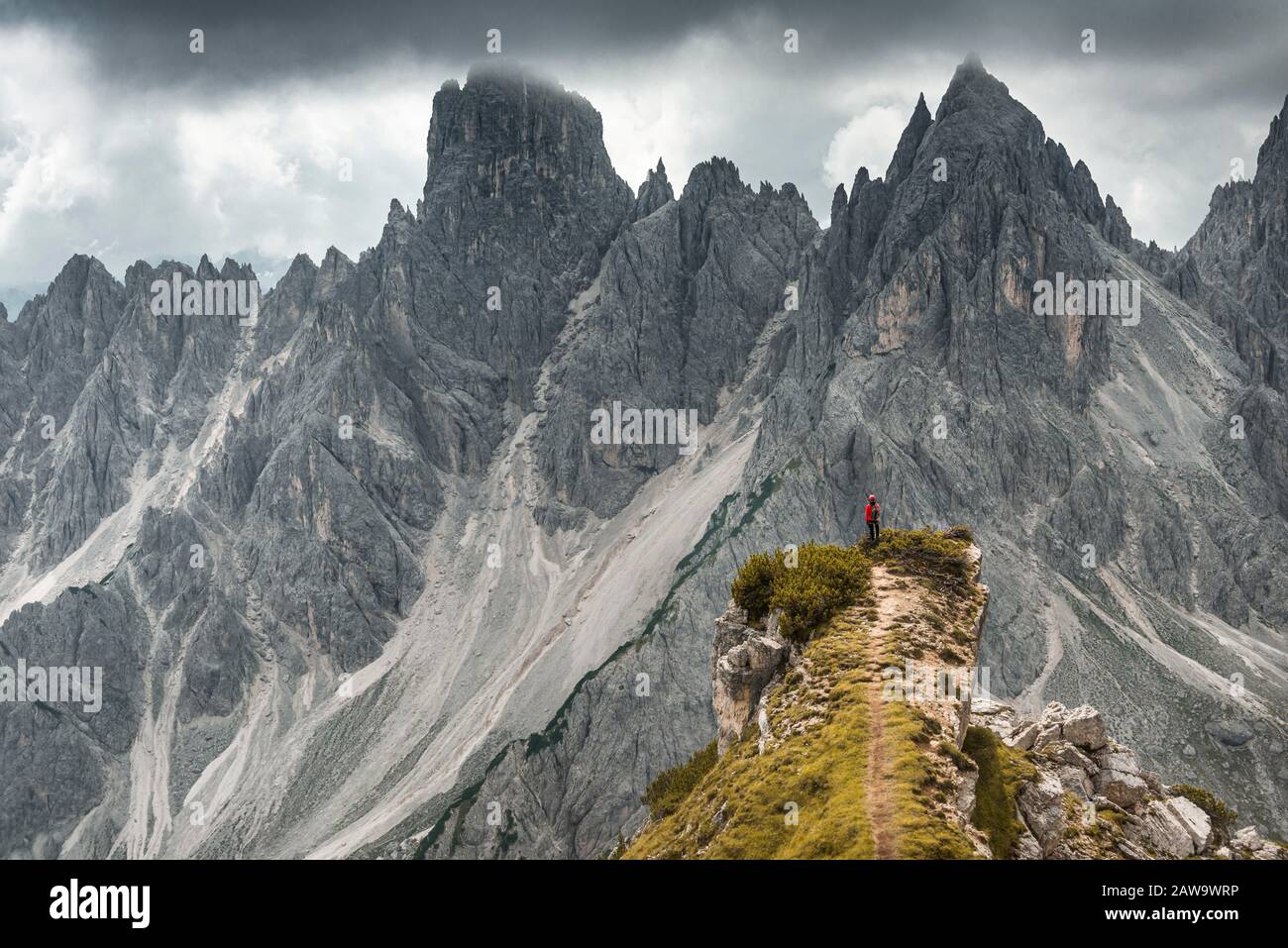 Homme avec veste rouge debout sur un degré, derrière lui des pics de montagne et des roches pointues, des nuages dramatiques, Cimon le groupe Croda Liscia et Cadini Banque D'Images