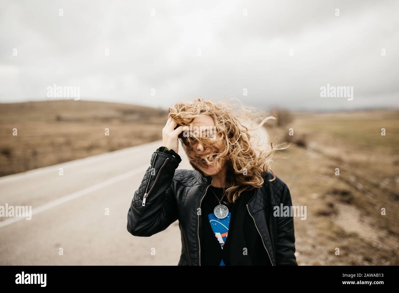 Portrait de la jeune femme avec des cheveux soufflant dans le vent. Elle porte une veste en cuir avec une nature et une route floues en arrière-plan. Banque D'Images