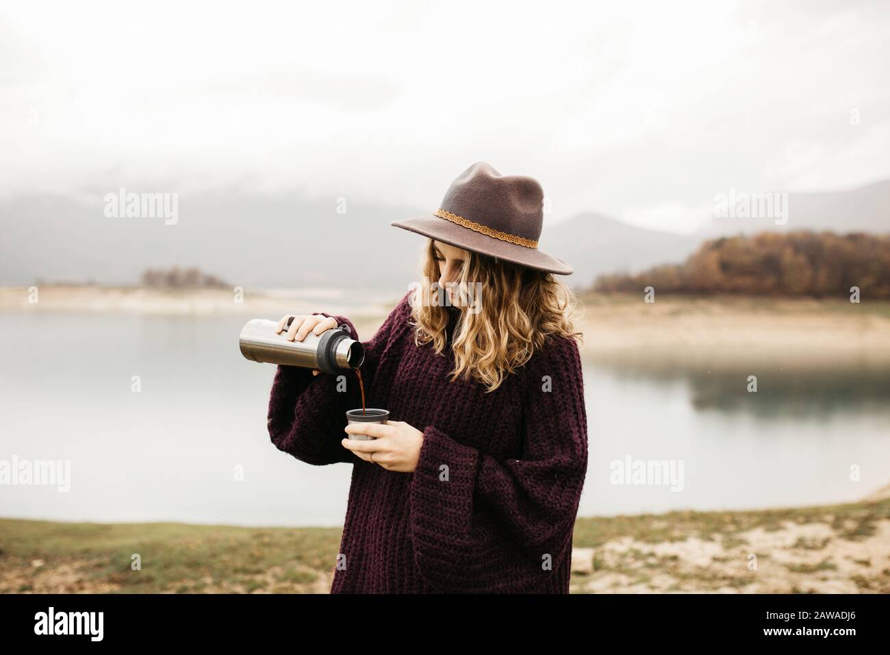 Heureuse belle femme avec chapeau buvant du café dans la nature, sur un lac de matin brouillard et les montagnes sont en arrière-plan. Elle sourit et déporte le café Banque D'Images