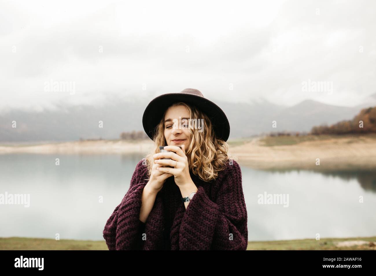 Heureuse belle femme avec chapeau buvant du café dans la nature, un matin brumeux. Le lac et les montagnes sont en arrière-plan. Banque D'Images
