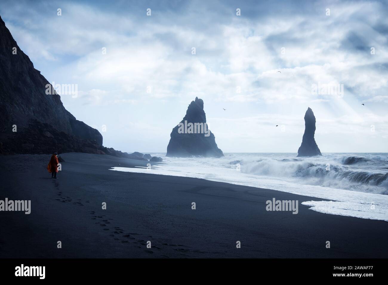 Voyageur masculin seul se tenant sur la plage de sable noir en Islande en regardant les vagues et les roches avec le soleil brillant à travers les nuages Banque D'Images