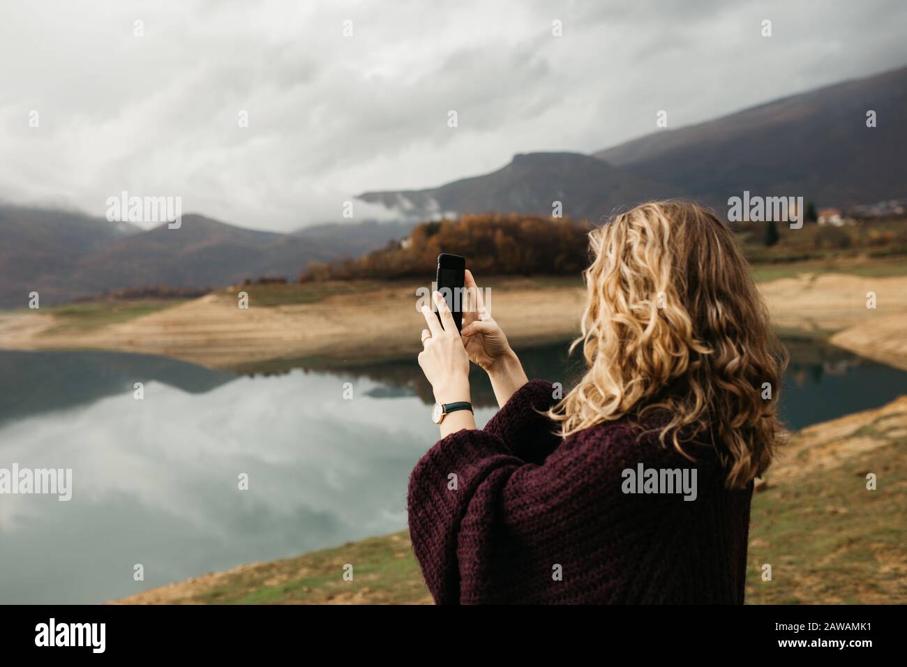 Belle femme avec des cheveux bouclés tenant le téléphone mobile et de prendre des photos du lac sur un jour nuageux. Elle envoie des SMS sur son smartphone et prend son selfie. Banque D'Images
