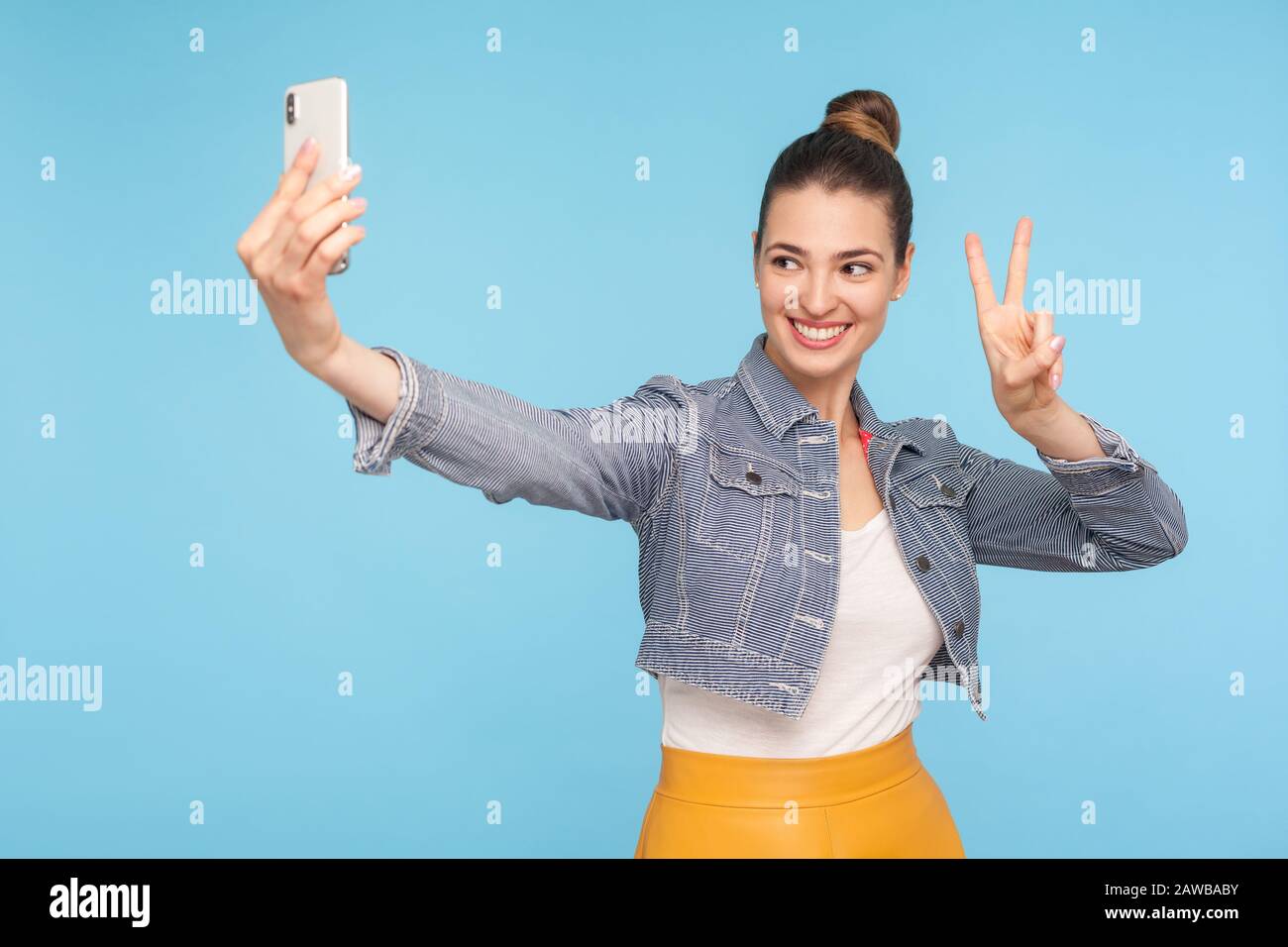 Jolie femme heureuse avec un petit pain dans un look tendance montrant la victoire ou le geste de paix tout en communiquant par appel vidéo, en prenant selfie et gestu Banque D'Images