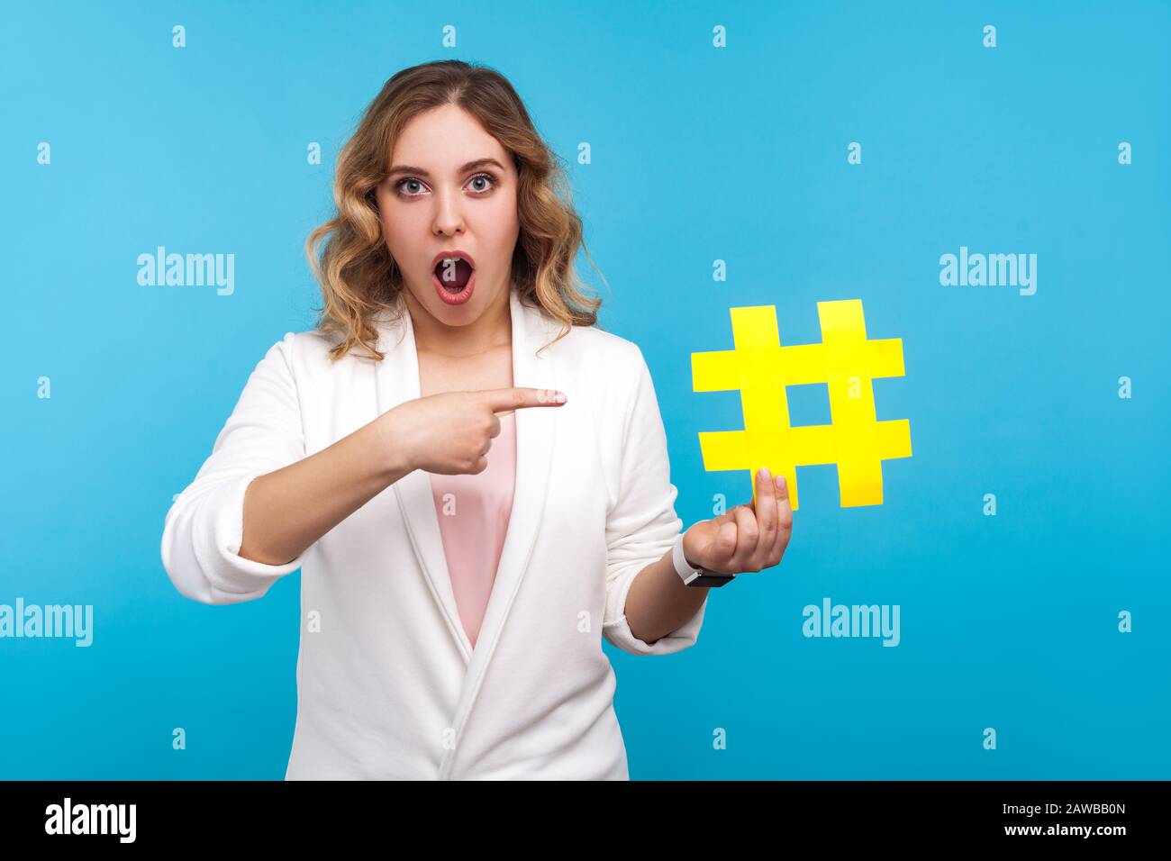 Des tendances multimédias incroyables. Portrait de blogger choqué, belle femme avec des cheveux ondulés dans la veste blanche pointant sur le symbole de hashtag de grand papier, regardant un Banque D'Images