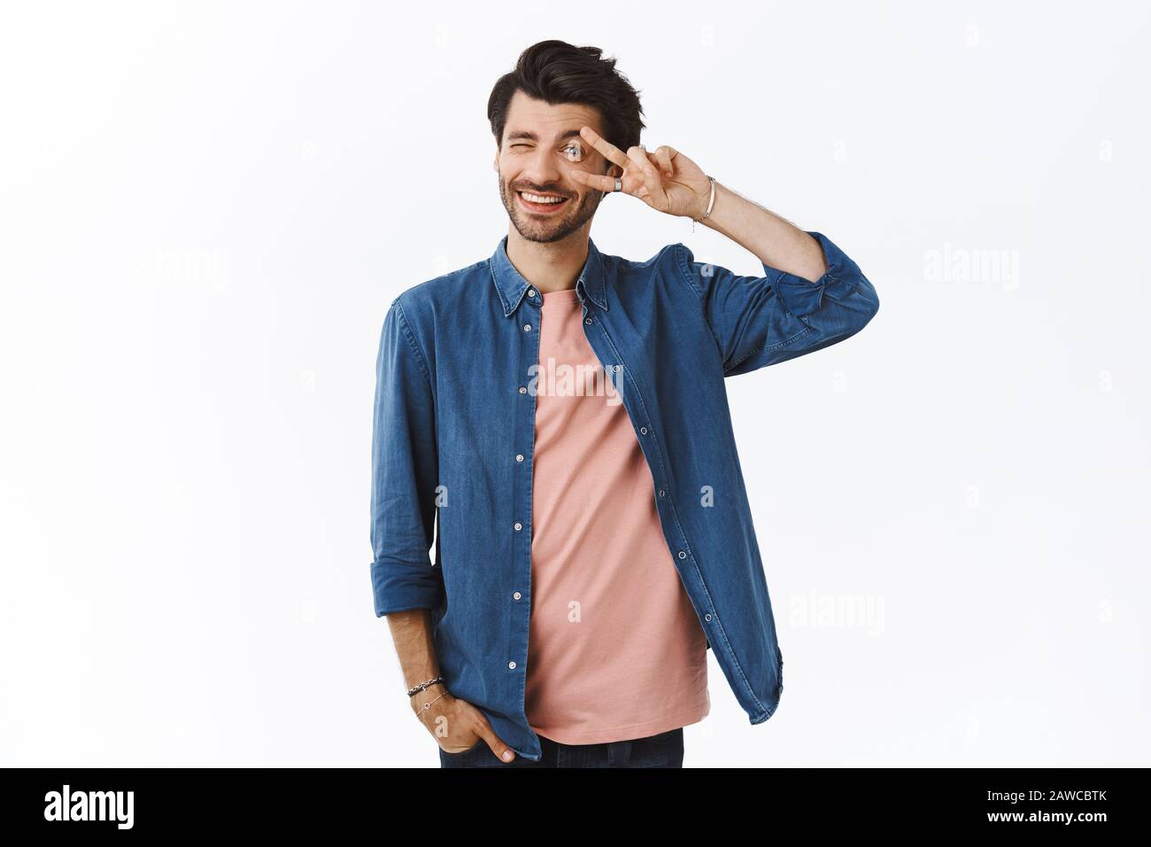 Joyeux petit ami à la barbe, mignon et soyeux, montrant la paix ou la victoire signe sur les yeux, à la chageuse à l'appareil photo et donner un sourire adorable, tenir une main dedans Banque D'Images