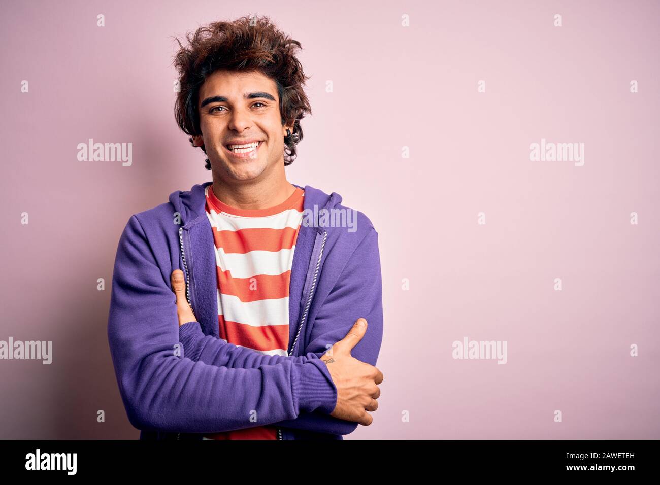 Jeune homme portant un t-shirt rayé et un sueur violet sur un arrière-plan rose isolé heureux visage souriant avec des bras croisés regardant l'appareil photo. Position Banque D'Images