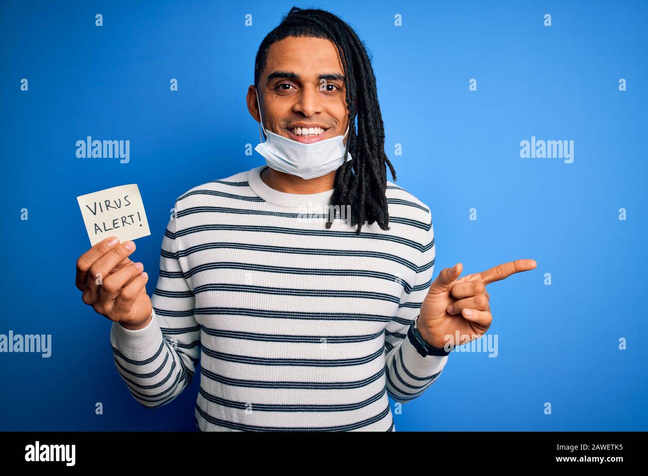 Un homme africain américain avec des cadenas portant un masque médical  tenant un rappel d'alerte de virus très heureux pointant avec la main et le  doigt sur le côté Photo Stock -