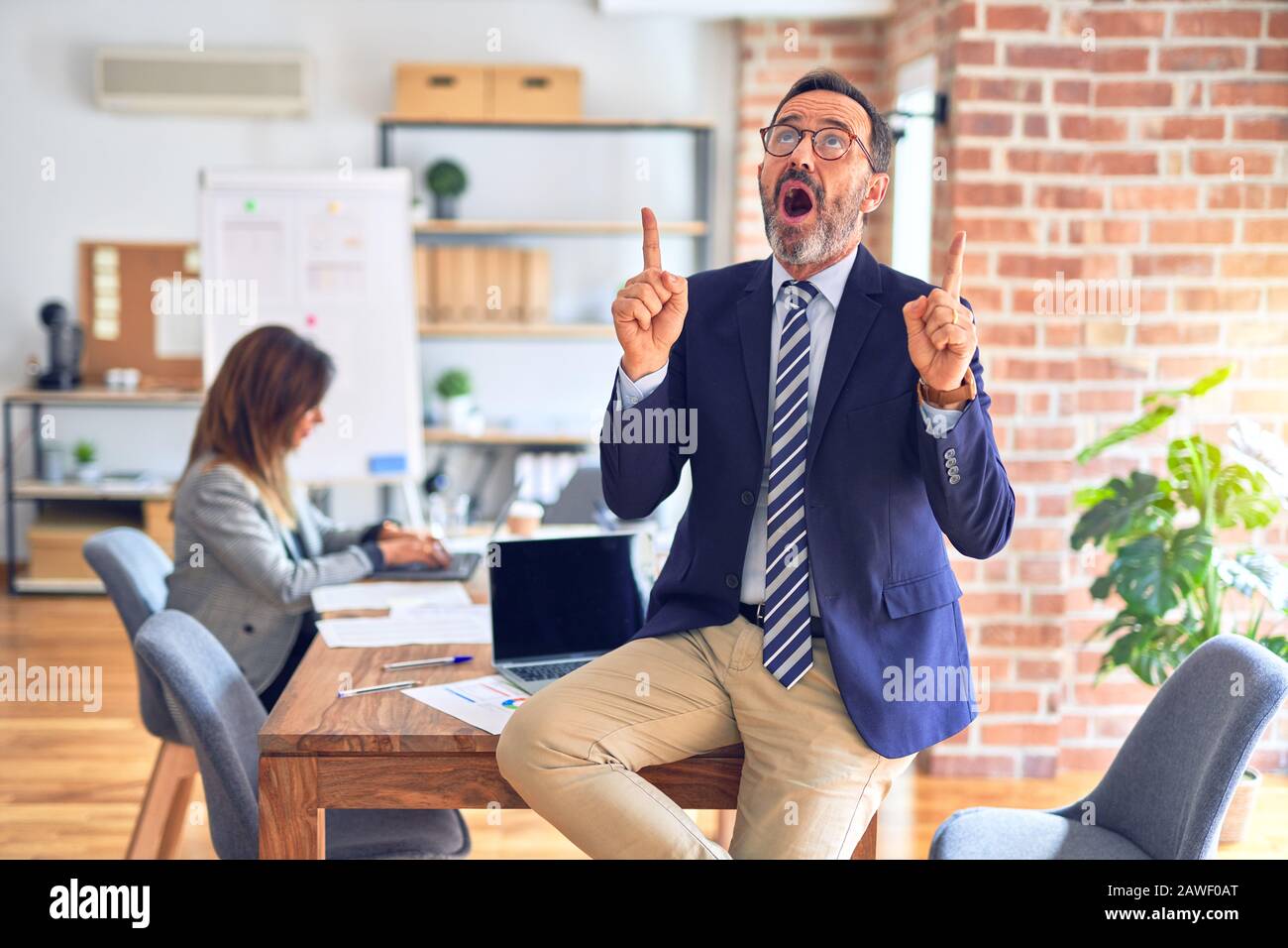 Homme d'affaires en âge moyen portant des lunettes assis sur le bureau étonné et surpris de regarder et de pointer avec les doigts et le bras levé Banque D'Images