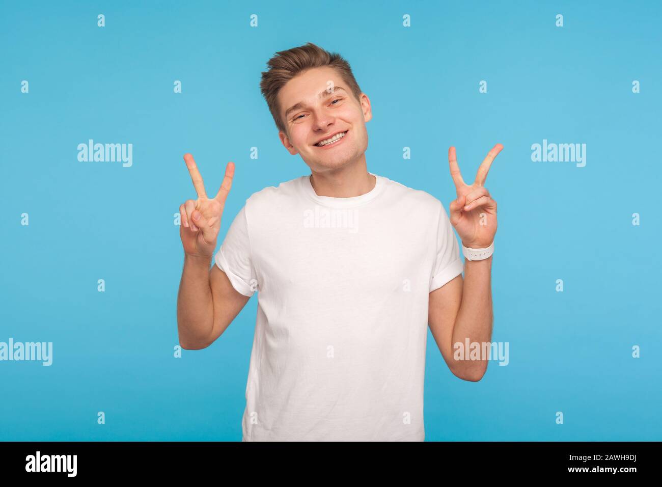 Portrait de l'homme heureux enchanté dans un t-shirt blanc montrant le signe de victoire avec les doigts doubles, gestant la paix et regardant l'appareil photo avec le sourire amical. Banque D'Images