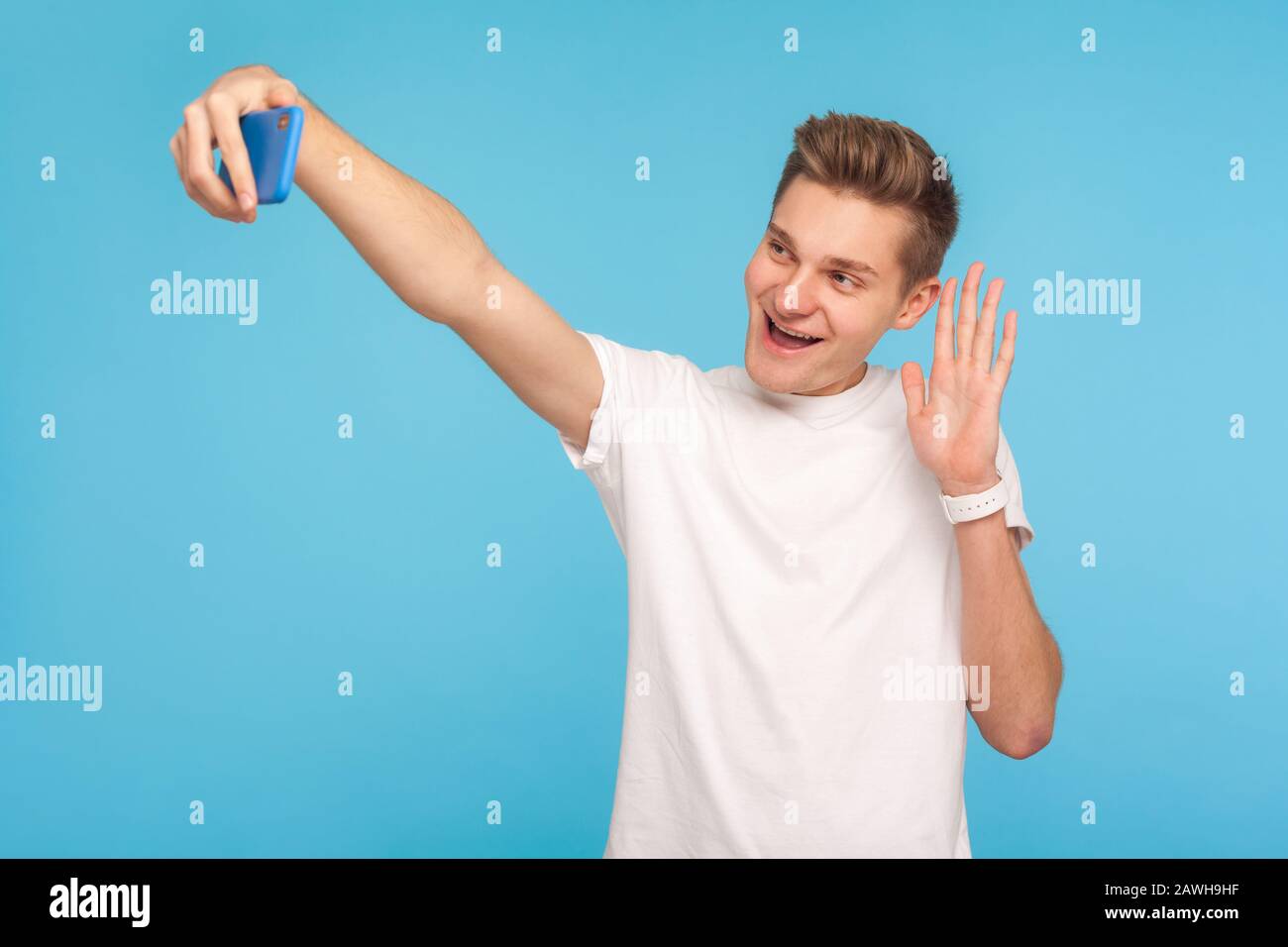 Sympathique heureux homme dans un t-shirt blanc décontracté gestuelle bonjour, dire hi tout en parlant sur appel vidéo, faire selfie à l'aide du téléphone portable, chat en ligne. Banque D'Images