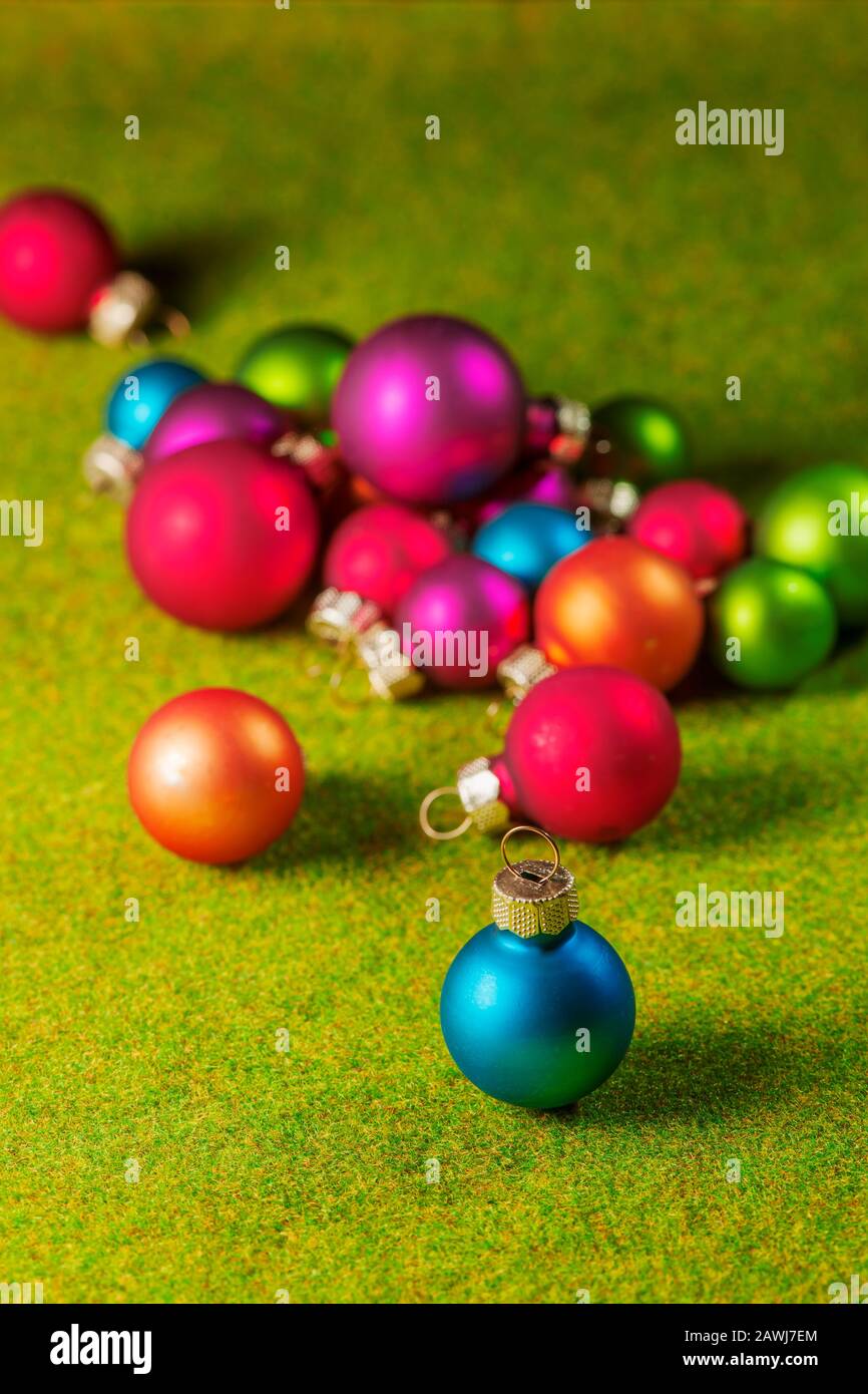Petites boules de Noël colorées floues sur l'herbe artificielle verte avec boule de Noël bleue sur pied unique au premier plan. Banque D'Images