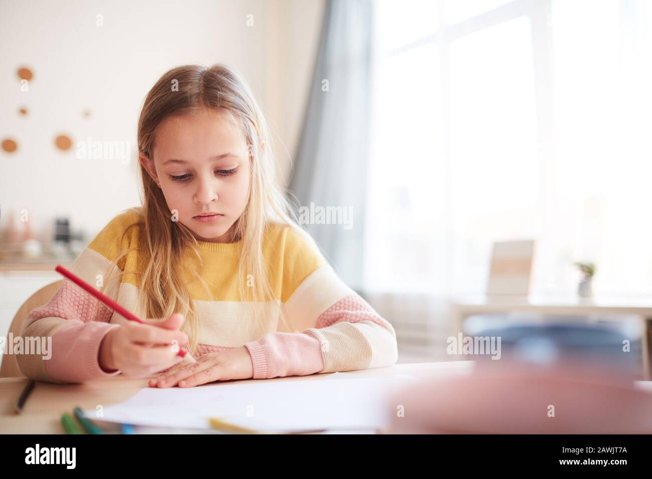 Portrait aux tons chauds de la jolie petite fille dessinant des images ou faisant des devoirs tout en étant assis à la table dans l'intérieur de la maison, espace de copie Banque D'Images