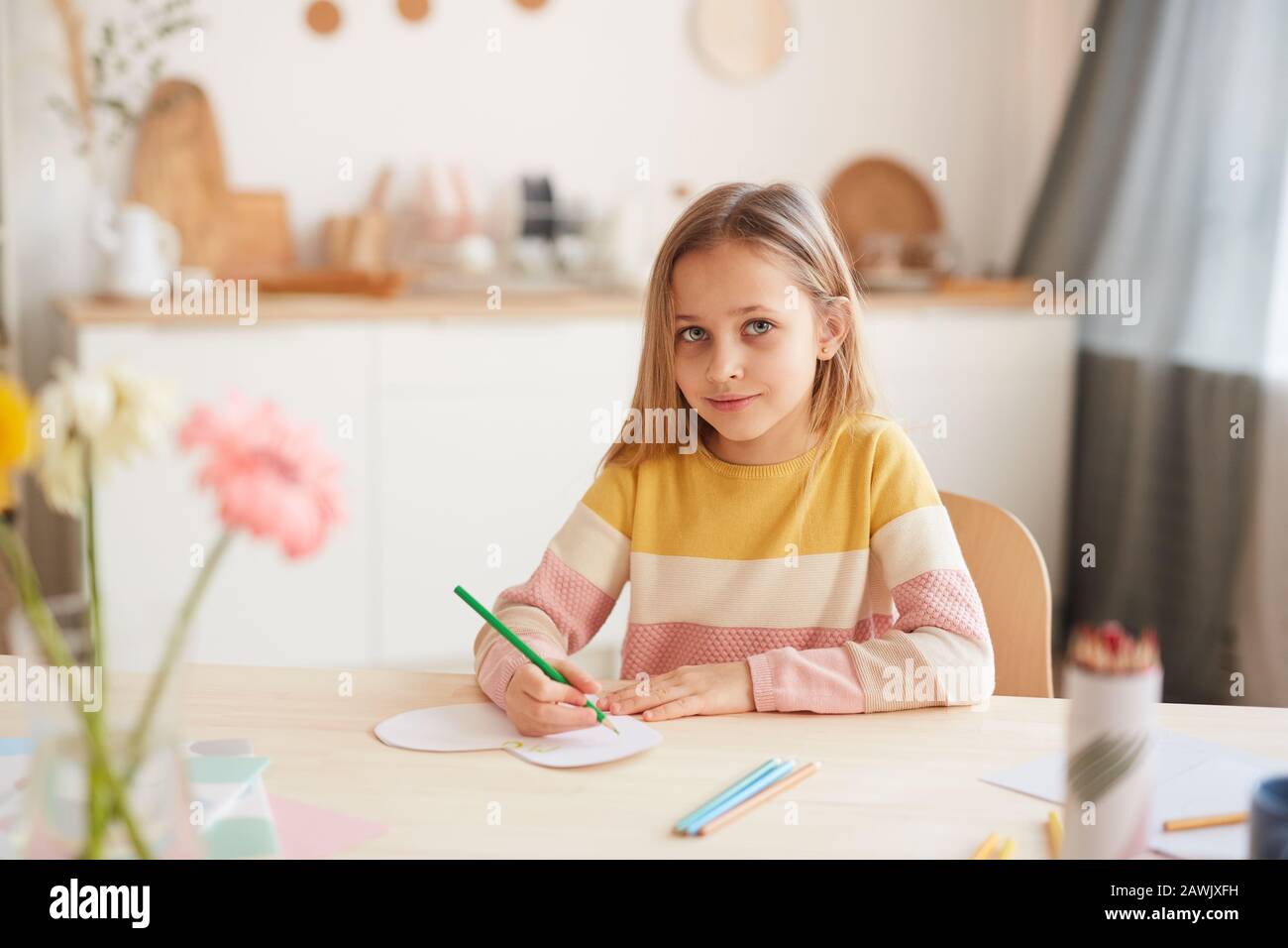 Portrait aux tons chauds de la petite fille mignonne regardant l'appareil photo et souriant tout en dessinant des photos ou en faisant des devoirs à la table à l'intérieur de la maison, espace de copie Banque D'Images