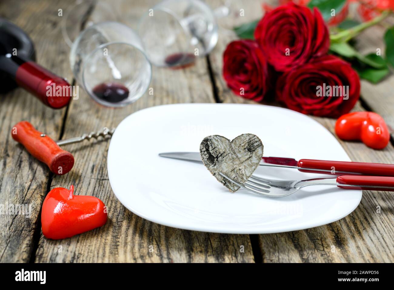 Coeur en bois sur une assiette avec fourche et couteau а, bouteille de vin а, corkvec, deux vitivinicoles, roses rouges et bougies sur une vieille table en bois. Mise au point sélective Banque D'Images