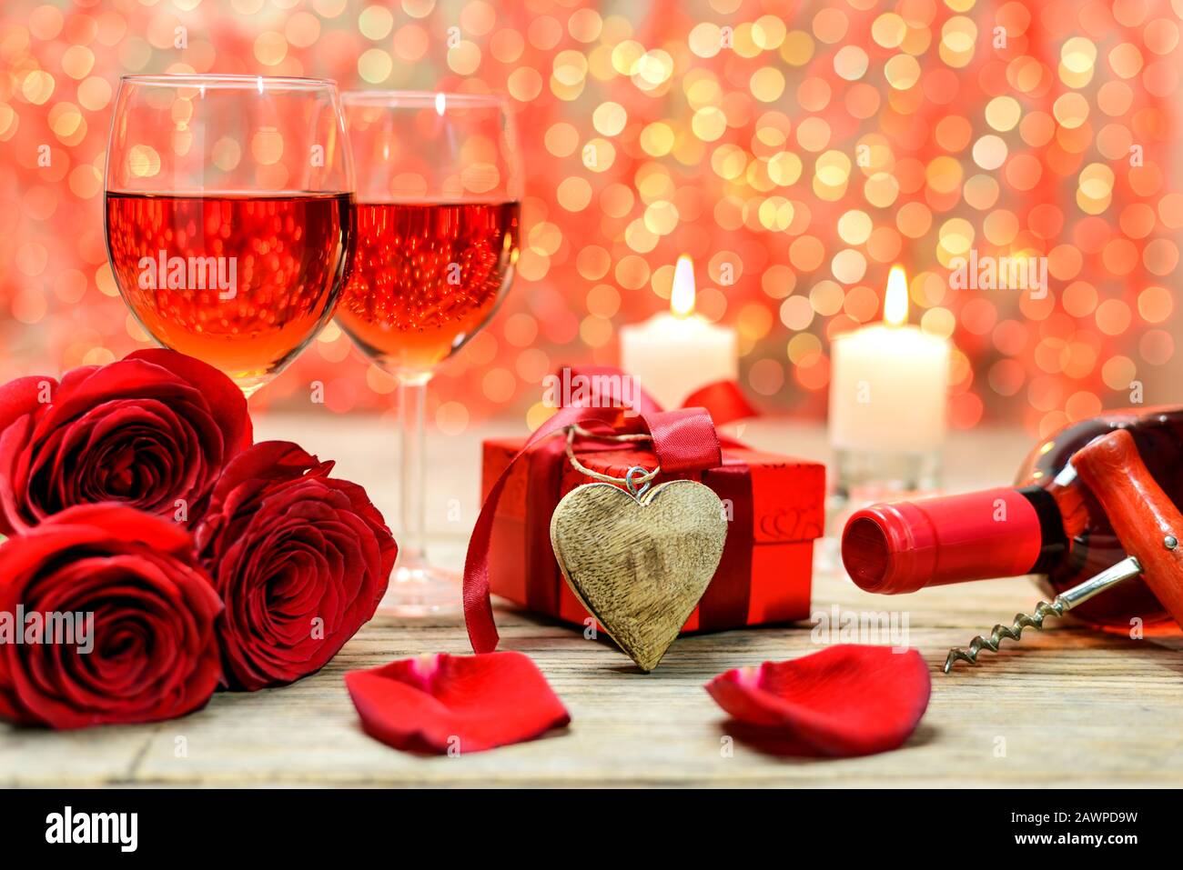 Coeur en bois, roses rouges, boîte à cadeaux, bouteille de vin, corkscrew, verres de vin et bougies brûlantes sur une vieille table en bois devant une lumière floue Banque D'Images