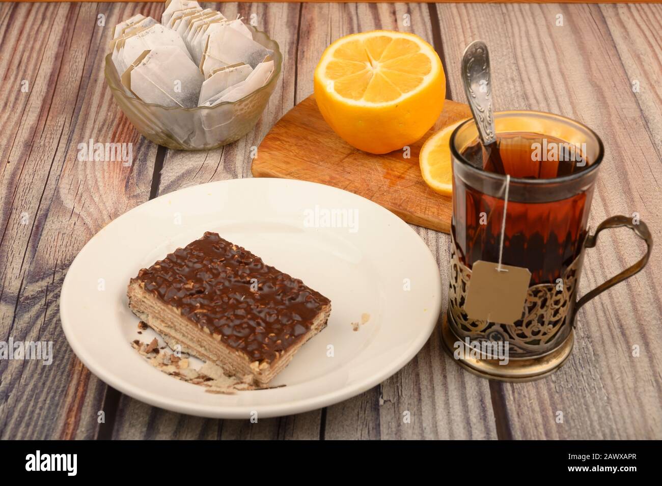 Thé dans un verre à facettes dans un porte-gobelet millésime, sachets de thé dans un vase en verre, citron tranché sur un plateau et un morceau de gâteau à gaufres sur une plaque blanche sur un woo Banque D'Images