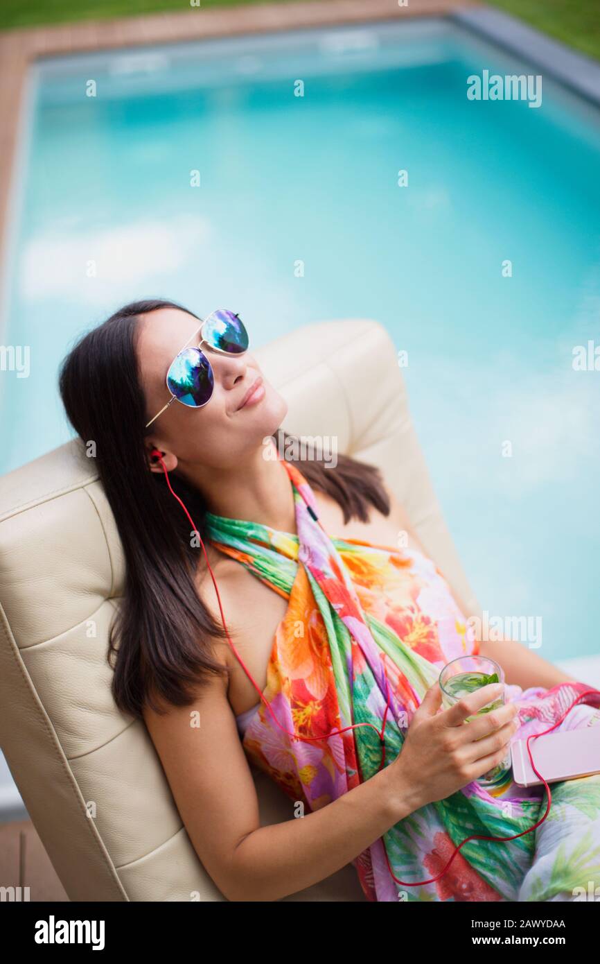 Femme sereine avec casque et cocktail relaxant, à l'écoute de la musique au bord de la piscine Banque D'Images
