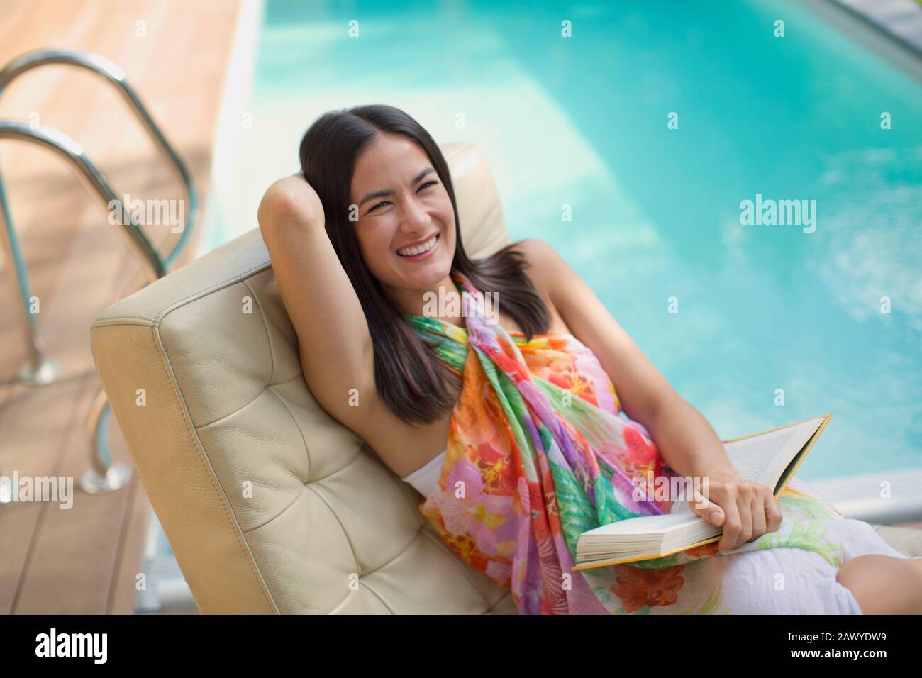 Portrait heureux, rire femme lecture livre sur chaise de salon à la piscine d'été Banque D'Images