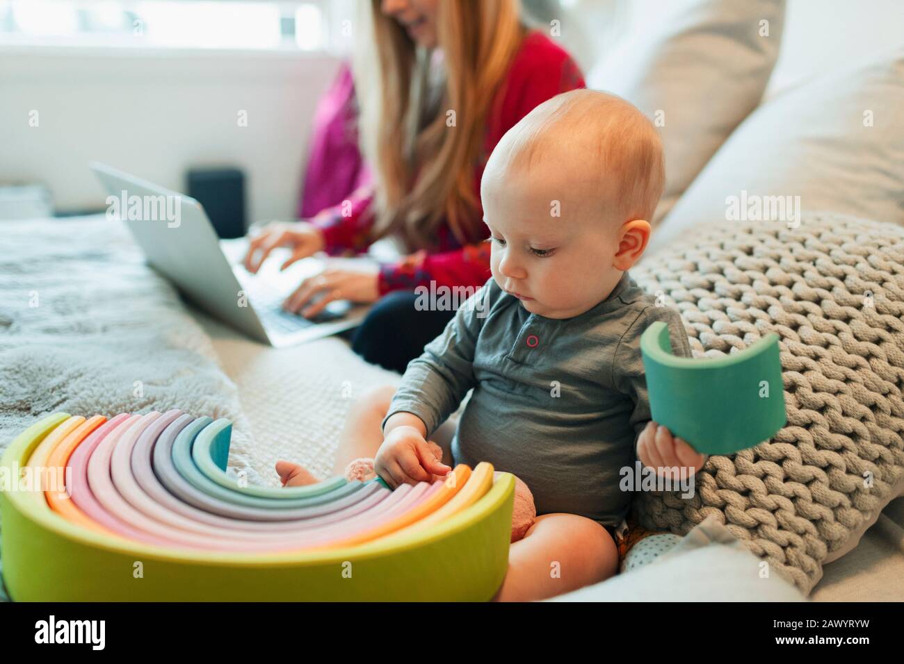 Curieux fille de bébé jouant avec le jouet sur le lit alors que la mère travaille à l'ordinateur portable Banque D'Images