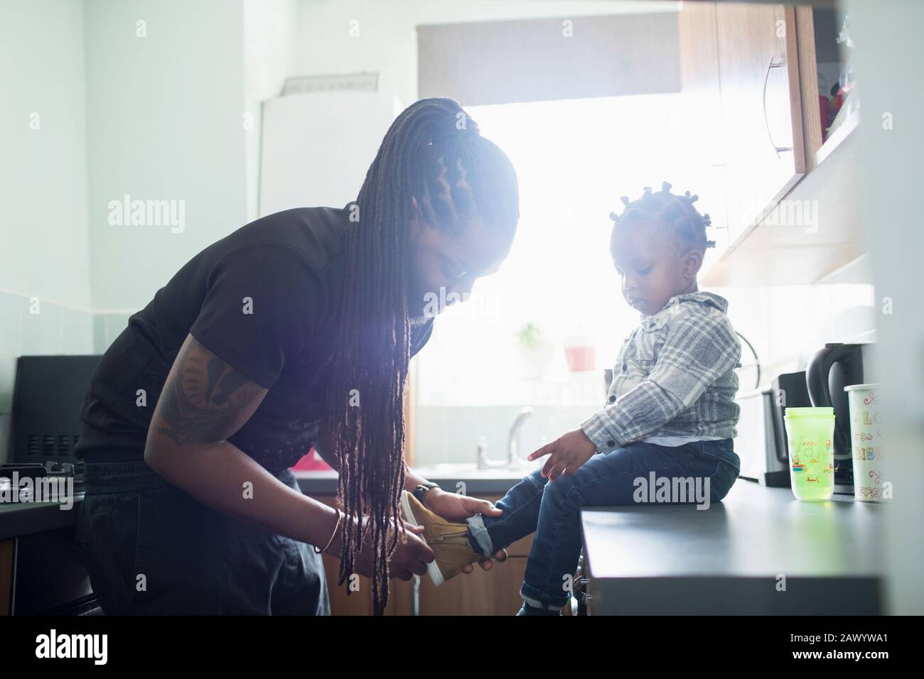 Père avec de longues tresses mettant des chaussures sur le petit enfant dans la cuisine ensoleillée Banque D'Images