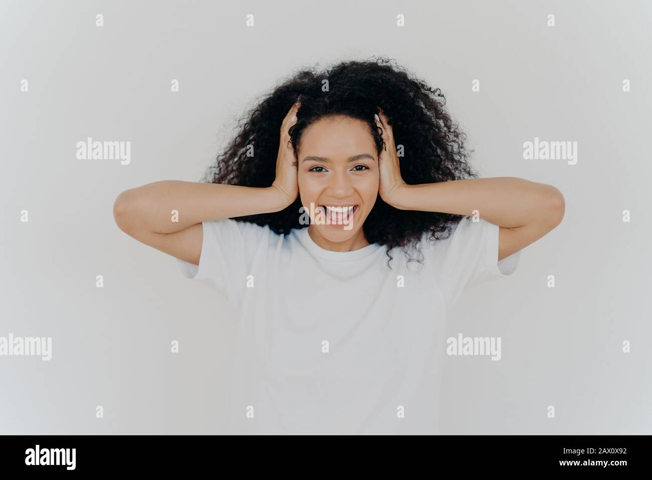 Photo d'une femme sombre et joyeuse à peau foncée garde les mains sur la tête, les cheveux frisés, les rires de joie, excitée par des émotions positives, vêtu d'un t-shirt blanc, entend f Banque D'Images